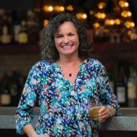 A woman standing in front of a bar holding a glass.